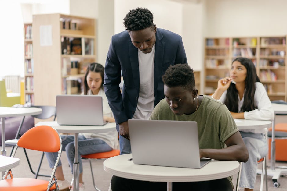 Students learning in a classroom setting with a teacher assisting and laptops on desks, creating an interactive education environment