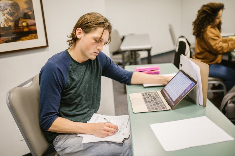 College student writing notes and using a laptop at a classroom desk for exam preparation